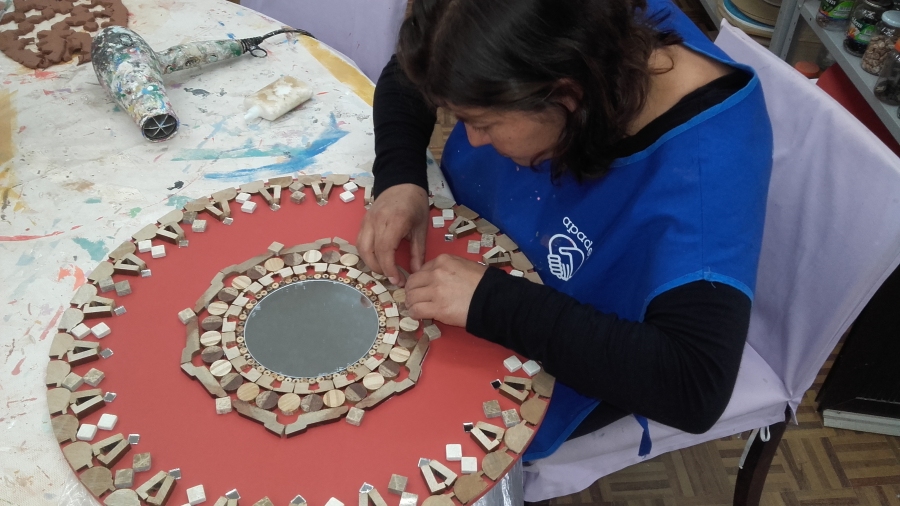 Imagem: Usuária sentada à mesa confeccionando uma Mandala redonda, vermelha, com peças de madeira. 